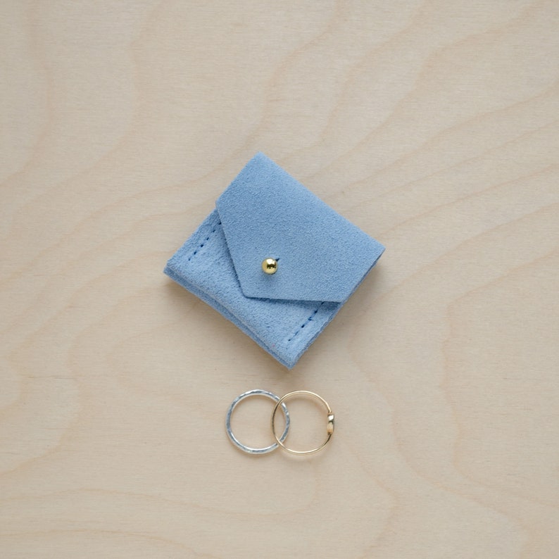 May include: A light blue suede ring box with a gold button closure. Two rings, one silver and one gold, are sitting on a wooden surface.
