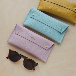 May include: A collection of leather eyeglass cases in pastel colors: lavender, light blue, and yellow. Each case has a flap closure with a gold stud and the lavender case is personalized with the name "SOPH". A pair of tortoiseshell sunglasses rests in front.