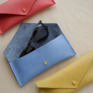 Envelope Glasses case in poppy red, powder blue and mustard yellow sit on wooden table. The blue one in the middle is open and shows a pair of tortoiseshell glasses inside.