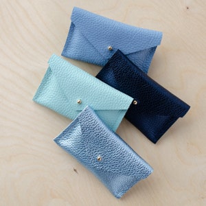 May include: Four leather wallets in different colours: light blue, teal, dark blue, and silver. The wallets are all closed and have a flap closure.