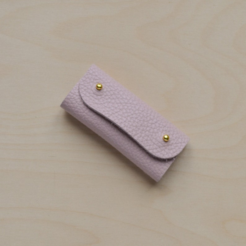 May include: A small, light pink leather key holder with gold accents. The key holder has a flap that folds over to secure keys.