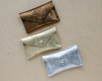 May include: Three small, metallic envelope-style wallets. The wallets are made of a shimmery material and are in gold, silver, and bronze.
