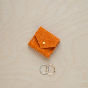 May include: A small orange suede pouch with a gold button closure. Two silver and gold rings are sitting next to the pouch.