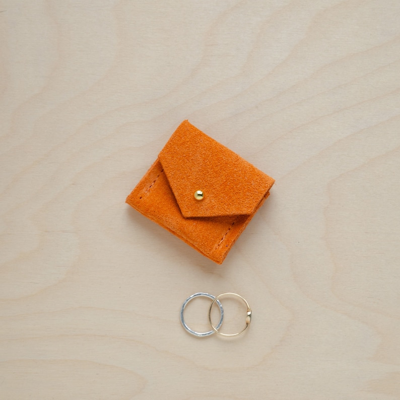 May include: A small orange suede pouch with a gold button closure. Two silver and gold rings are sitting next to the pouch.