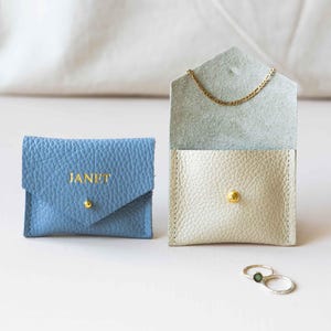 May include: A blue leather envelope-style pouch with the name JANET embossed in gold, alongside an open cream-coloured leather pouch containing a gold chain necklace and two silver rings with a small green stone resting nearby.