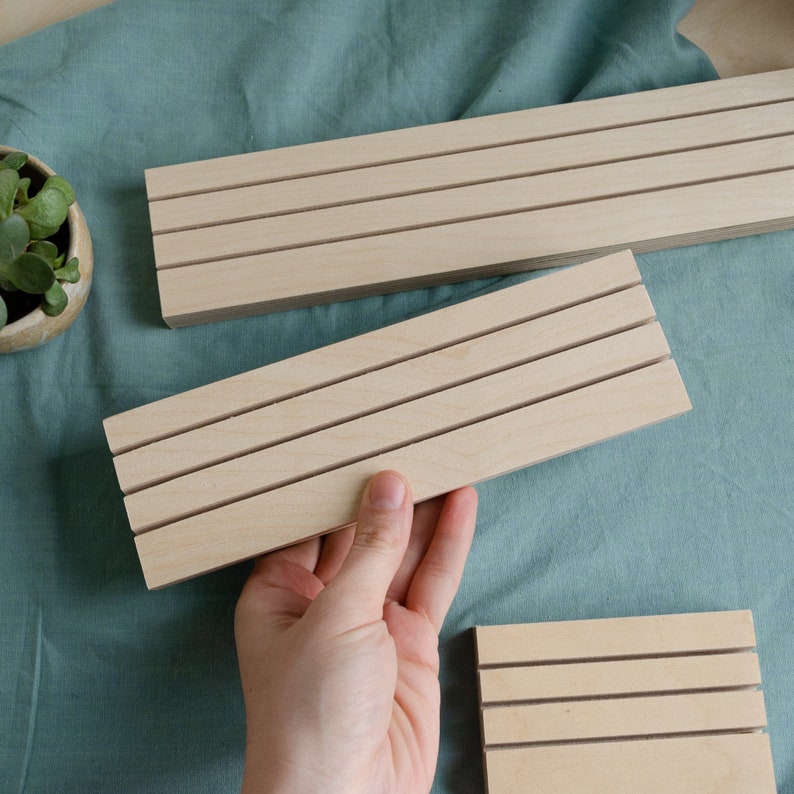 K&ouml;nnte beinhalten: Mehrere unfertige Holz-Ausstellungsst&auml;nder mit horizontalen Rillen. Ein St&auml;nder wird von einer Hand gehalten. Die St&auml;nder sind rechteckig und aus hellem Holz gefertigt. Der Hintergrund ist ein t&uuml;rkisfarbener Stoff.