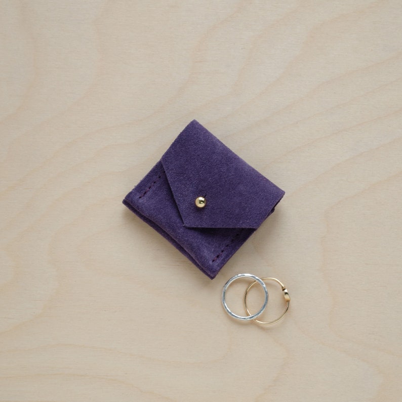 May include: A small purple suede pouch with a gold button closure. Two rings, one gold and one silver, are sitting next to the pouch.
