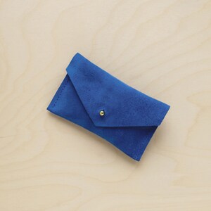 May include: A vibrant blue suede envelope-style wallet with a gold-colored snap closure. The wallet is rectangular with a triangular flap. The background is a light wood grain.
