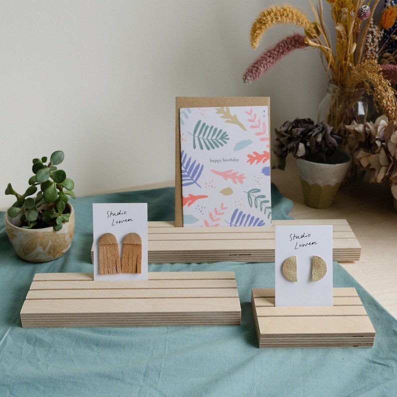 K&ouml;nnte beinhalten: Pr&auml;sentation von handgefertigten Ohrringen und einer Gru&szlig;karte auf Holzplattformen. Die Ohrringe haben beige Fransen und goldene Halbkreis-Designs. Die Karte hat ein Blumenmuster und tr&auml;gt die Aufschrift "happy birthday".