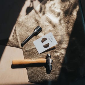 May include: A metal hammer with a wooden handle, a metal tool, and a white card with the text "Studio Lumen" and a design of two half-circles, are arranged on a textured, gold-colored surface.