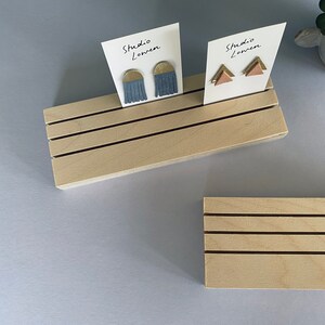 K&ouml;nnte beinhalten: Zwei helle Holzst&auml;nder mit horizontalen Rillen, die jeweils wei&szlig;e Karten mit der Aufschrift Studio Lowen halten. Eine Karte zeigt blaue halbkreisf&ouml;rmige Fransenohrringe, die andere zwei kleine rosa dreieckige Ohrringe.