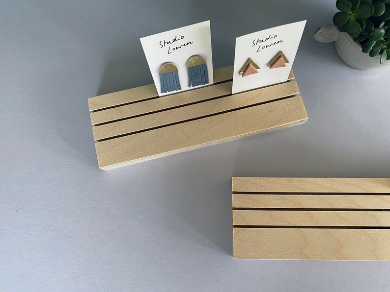 K&ouml;nnte beinhalten: Zwei helle Holzst&auml;nder mit horizontalen Rillen, die jeweils wei&szlig;e Karten mit der Aufschrift Studio Lowen halten. Eine Karte zeigt blaue halbkreisf&ouml;rmige Fransenohrringe, die andere zwei kleine rosa dreieckige Ohrringe.