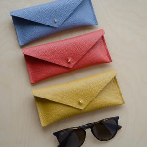 Envelope Glasses case in poppy red, powder blue and mustard yellow sit on wooden table alongside a pair or sunglasses. The eyeglasses cases have a minimalist design featuring an envelope style. The flap is secured with a simple stud fastening.