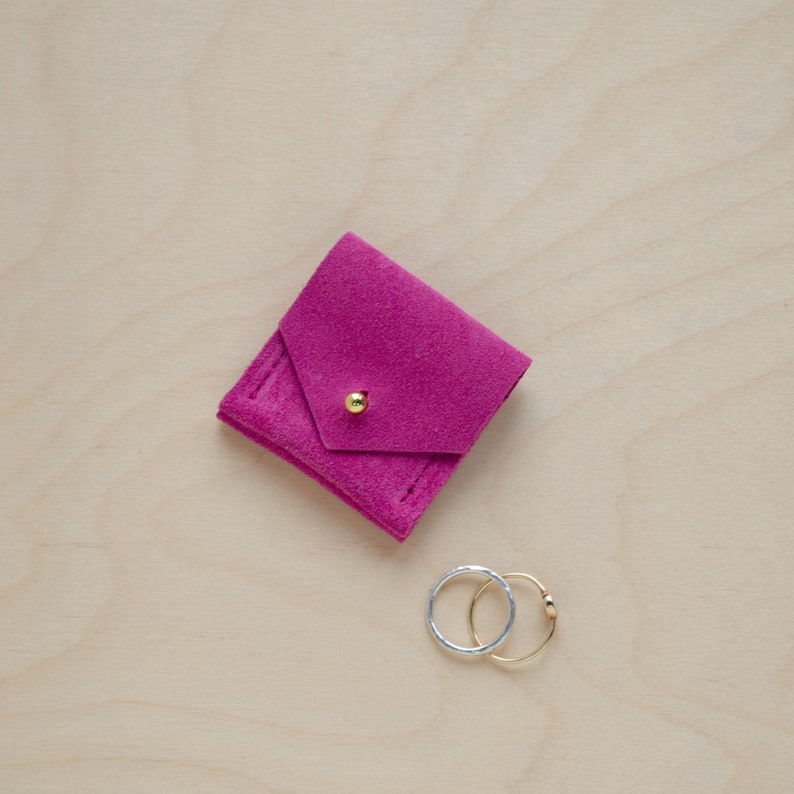 May include: A small pink suede pouch with a gold button closure. Two rings, one silver and one gold, are sitting next to the pouch.