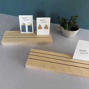 K&ouml;nnte beinhalten: Holz-Ausstellungsst&auml;nde mit schwarzen Linien halten Ohrringkarten. Eine Karte zeigt blaue Quastenohrringe mit goldenen Akzenten, die andere rosa Dreiecks-Ohrringe mit goldenen Akzenten. Der Text "Studio Lowen" steht auf den Karten.
