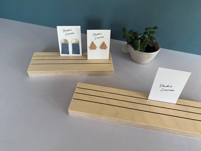 K&ouml;nnte beinhalten: Holz-Ausstellungsst&auml;nde mit schwarzen Linien halten Ohrringkarten. Eine Karte zeigt blaue Quastenohrringe mit goldenen Akzenten, die andere rosa Dreiecks-Ohrringe mit goldenen Akzenten. Der Text "Studio Lowen" steht auf den Karten.