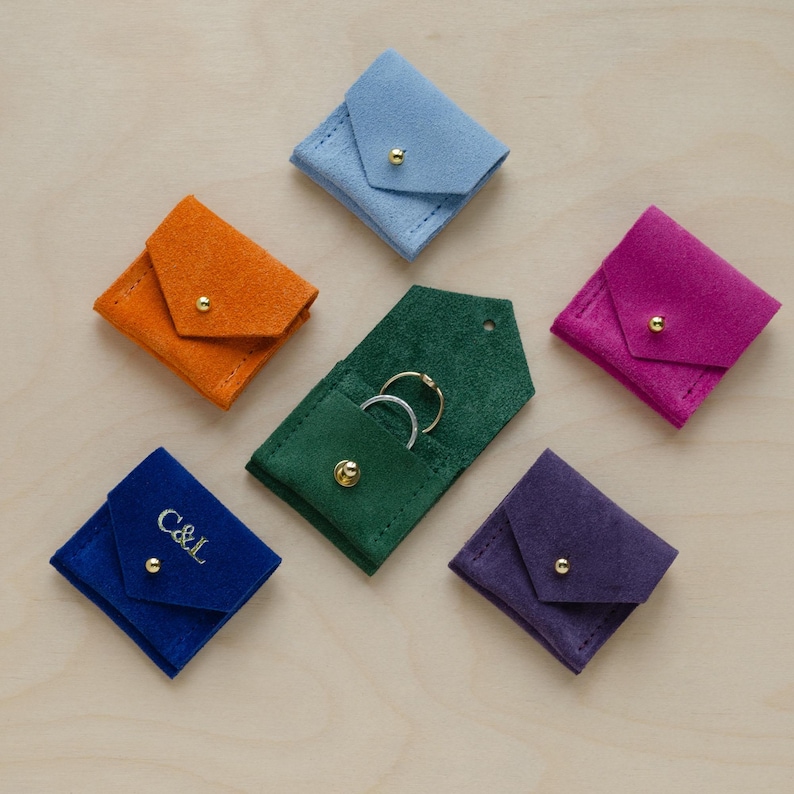 Six wedding ring pouches in suede. Colours include, Clementine Orange, Pale Blue, Ultramarine Blue, Moss Green, Grape Purple and Fuchsia Pink.

The Blue pouch is customised with personalisation of the bride and grooms initials in gold embossing.