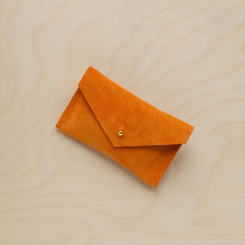 May include: An orange suede envelope-style wallet with a gold-colored stud closure. The wallet is rectangular with a folded flap and visible stitching along the edges. The background is a light wood grain.