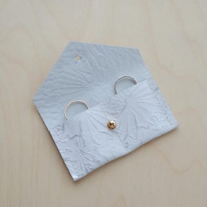 May include: A light grey, floral-patterned leather ring holder, open to display two rings. The holder is secured with a gold-coloured snap. The background is a light wood grain.