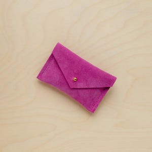 May include: A magenta suede envelope-style wallet with a gold-colored snap closure. The wallet is rectangular with a folded flap and visible stitching along the edges. The background is a light wood grain.