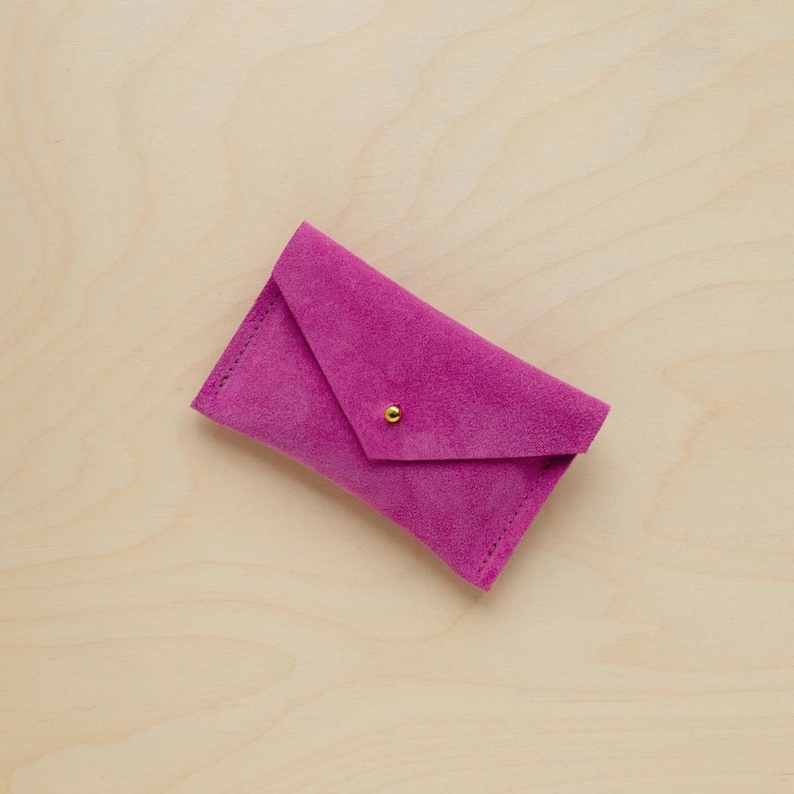 May include: A magenta suede envelope-style wallet with a gold-colored snap closure. The wallet is rectangular with a folded flap and visible stitching along the edges. The background is a light wood grain.