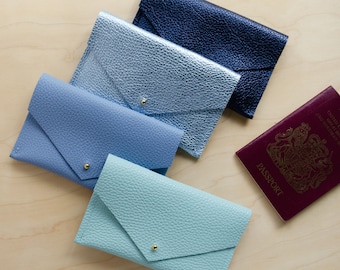 May include: Four leather passport holders in different colors: light blue, blue, silver, and light green. The holders are arranged in a stack, with the light blue holder on top. A burgundy passport with the word "PASSPORT" printed on the cover is lying on a wooden surface.