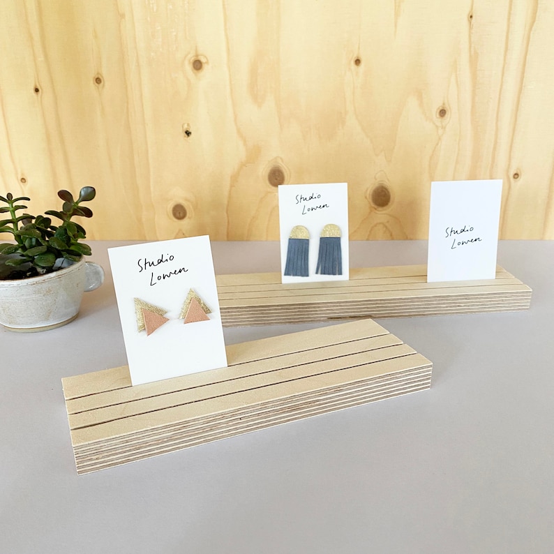 K&ouml;nnte beinhalten: Handgefertigte Ohrringe auf Holzst&auml;ndern. Die Ohrringe werden auf wei&szlig;en Karten mit dem Text "Studio Lowen" pr&auml;sentiert. Die Ohrringe haben Dreiecks- und Fransen-Designs in verschiedenen Farben.