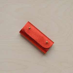 May include: A small, red leather key holder with gold accents. The key holder is made of a textured leather and has a flap that closes with a snap.