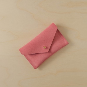 May include: A dusty rose-colored suede envelope-style wallet with a gold-colored button closure. The wallet is rectangular with a triangular flap and visible stitching along the edges. The background is a light wood grain.