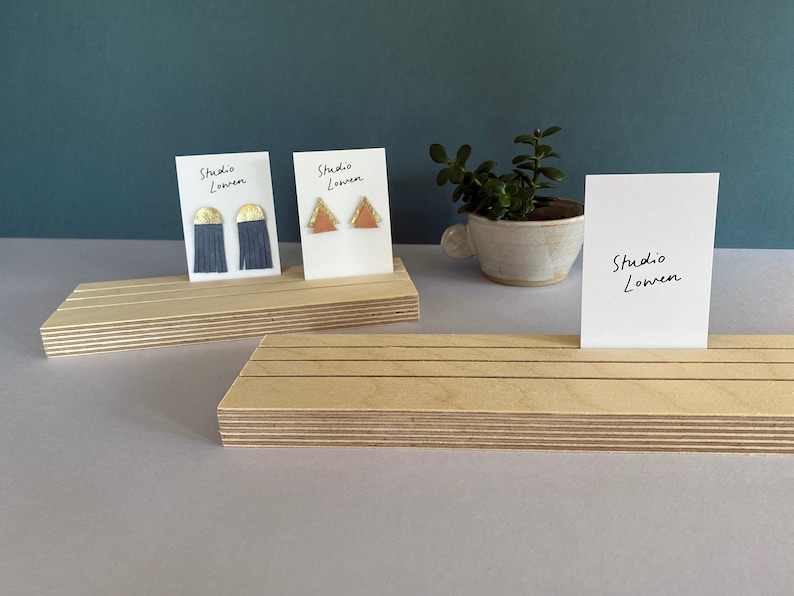 K&ouml;nnte beinhalten: Ohrringe auf Holzst&auml;ndern pr&auml;sentiert. Ein Paar hat marineblaue Fransen mit goldenen Akzenten, das andere orangefarbene Dreiecke mit goldenen Details. Die St&auml;nder halten Karten mit "Studio Lonren"-Schrift.