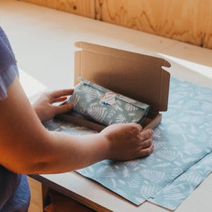 May include: A person opening a small, brown cardboard box containing a wrapped item. The item is wrapped in light green fabric with a white fern pattern. The wrapping paper and the surface it is on have the same fern pattern.
