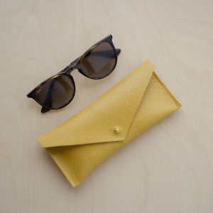 Yellow Leather Envelope Glasses case sits  on wooden table alongside a pair or sunglasses. The eyeglasses cases have a minimalist design featuring an envelope style. The flap is secured with a simple stud fastening.