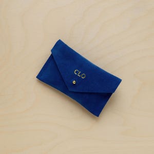 May include: A royal blue, folded envelope-style case with a gold snap closure. The case is made of a soft, velvety material and features the gold-colored initials "CLO" embroidered on the front.
