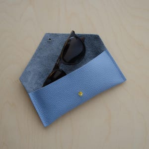 Blue Leather Envelope Glasses case sits  on wooden table alongside a pair or sunglasses. The eyeglasses cases have a minimalist design featuring an envelope style. The flap is secured with a simple stud fastening.