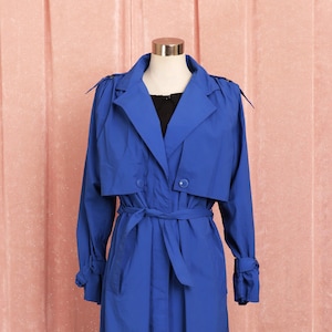May include: A long, royal blue trench coat with a belt and double breasted closure. The coat is on a mannequin against a pink background.