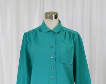 May include: A teal blue long-sleeved blouse with a collared neckline and a single pocket on the left chest. The blouse has a button-up front closure and a gathered detail at the shoulders.