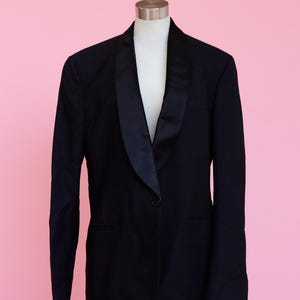 May include: A black blazer with a satin lapel. The blazer has a single button closure and is made of a smooth fabric.