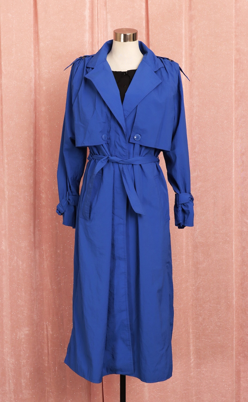 May include: A long, royal blue trench coat with a belt and double breasted closure. The coat is on a mannequin against a pink background.