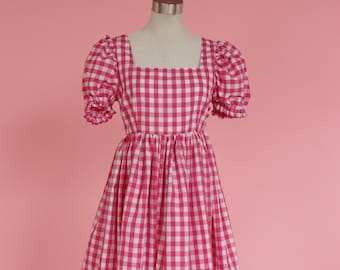 May include: A pink and white gingham dress with puffed sleeves and a square neckline. The dress is on a mannequin against a pink background.