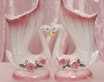 May include: A pair of white ceramic swan vases with pink and iridescent glaze. Each vase features a pink rose with green leaves on the side. The swans are facing each other with their necks curved to form a heart shape.
