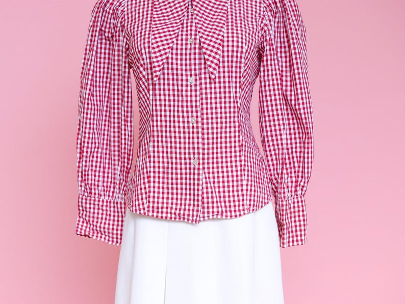 Puede incluir: Una camisa de botones de cuadros rojos y blancos con cuello Peter Pan y mangas largas. La camisa tiene una silueta ajustada y est&aacute; hecha de una tela ligera.