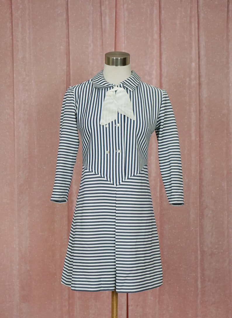May include: A white and navy blue striped dress with a white bow at the collar. The dress has a button-up front and a slightly flared skirt.