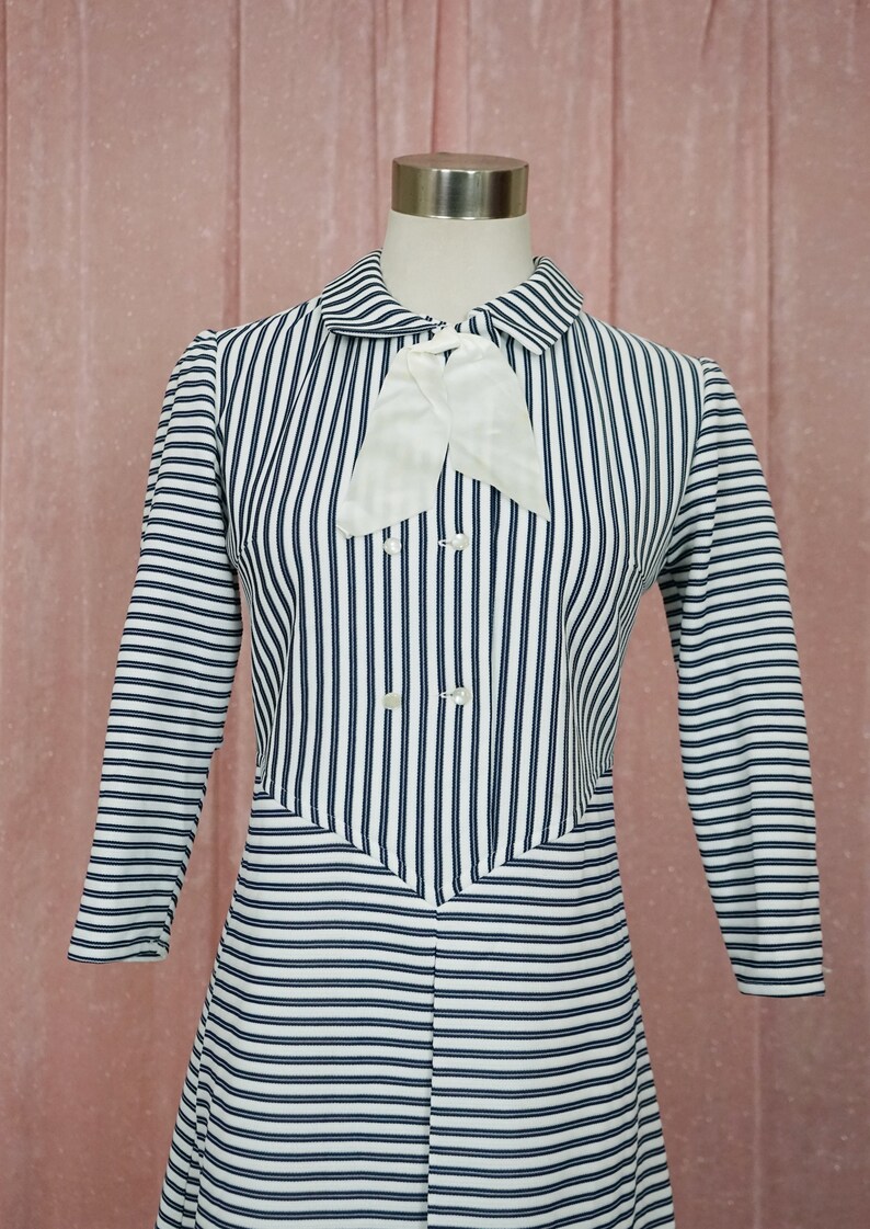 May include: A white and navy blue striped dress with a white bow at the collar. The dress has long sleeves and a button-up front.