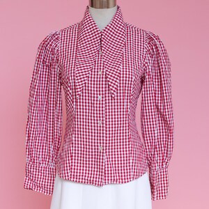 Puede incluir: Una camisa de botones de cuadros rojos y blancos con un cuello grande y puntiagudo. La camisa tiene mangas largas con hombros abullonados y una silueta ajustada.