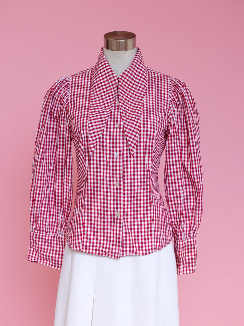 Puede incluir: Una camisa de botones de cuadros rojos y blancos con un cuello grande y puntiagudo. La camisa tiene mangas largas con hombros abullonados y una silueta ajustada.