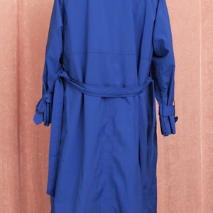 May include: A long, royal blue trench coat with a belt and long sleeves. The coat is on a mannequin against a pink background.