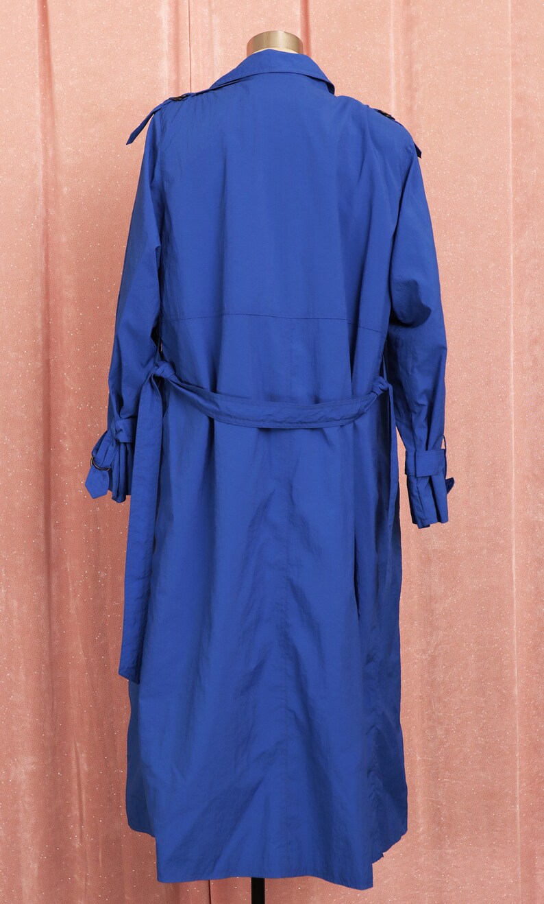 May include: A long, royal blue trench coat with a belt and long sleeves. The coat is on a mannequin against a pink background.