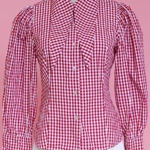 Puede incluir: Una camisa de botones de cuadros rojos y blancos con un cuello grande y puntiagudo. La camisa tiene mangas largas y una silueta ajustada.