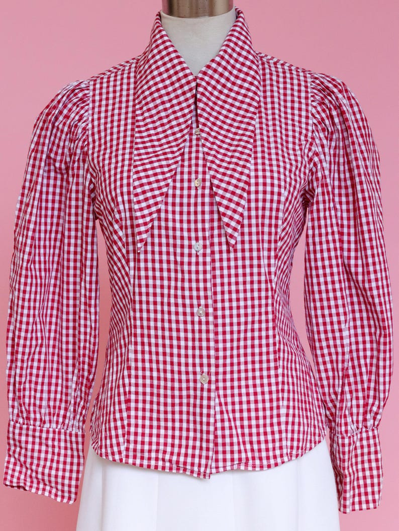 Puede incluir: Una camisa de botones de cuadros rojos y blancos con un cuello grande y puntiagudo. La camisa tiene mangas largas y una silueta ajustada.