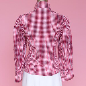 Puede incluir: Una camisa de botones de cuadros rojos y blancos con cuello alto y mangas largas. La camisa es entallada y tiene una manga ligeramente abullonada.